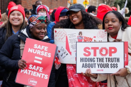 Three Black women nurses pose for camera; two are smiling, one looks determined. Two hold printed NYSNA signs: "Safe nurses = safe patients" and "STOP hiding the truth about your $$$" and the other holds a handwritten sign, partly covered, long text. Lots of red NYSNA hats are visible in the dense, upbeat crowd behind them.