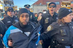 A man in a blue Amazon delivery driver jacket is flanked by bevy of cops, who are holding his arms and leading him away. More cops are behind him, and a crowd behind them. Printed Teamsters picket signs are visible saying "Amazon ULP strike" and "Amazon is unfair."