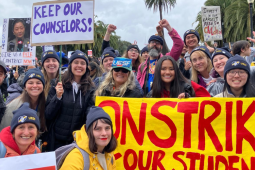 Smiling strikers pose outdoors with a bright yellow banner handpainted "On strike for our schools" in red. Most people visible in this photo appear to be white or Asian women. Many wear UESF hats. Other visible handmade signs say "Boo Su, give us a fair contract now!" "Keep our counselors" and "Invest in oiur largest asset: children." Palm trees are visible against gray sky. Some people raise fists or a victory/peace sign.