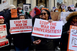 A big crowd, bundled against cold, stands in front of a building with columns. Foregrounded are four women, one with beaming smile, one looking up warmly at her. All hold PSC-CUNY signs that say variously, "Stop the War on Universities," "Save Research," and "Science is for the People." Other handmade signs, a UAW logo, and an upside-down American flag are visible in the crowd behind htem.