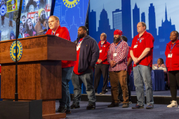 Autoworkers in red t-shirts stand on stage at a convention.