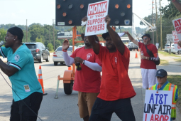 Black and white men, most in red union T-shirts, picket energetically on a streetcorner. Man in front, in turquoise shirt, is talking or chanting into a mic, arm outstretched. Everyone's pose is active. Printed signs say "honk if you support workers" and "AT&T unfair, CWA on strike, ULP." One handmade sign, partially visible, says "AT&T Unfair Labor." The man in the center is holding his sign high and wind or movement blows his red shirt.