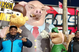 Two Amazon workers hold picket signs in front of an inflatable pig smoking a cigar and holding a bag of money.