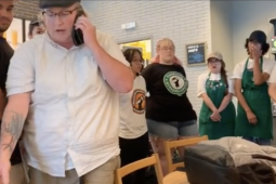 Starbucks workers in aprons surround a manager in a white shirt, the manager is leaving and talking on the phone