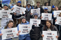 About 15 smiling people hold identical signs saying “ARISE for Higher Para Pay”