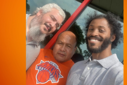 Three workers pose for a group selfie in front of the store. They read as three men of different races. The man in the middle is wearing an orange Knicks T-shirt.