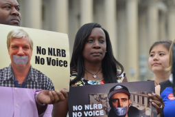 A man and a woman hold signs saying “no NLRB, no voice” with the columns of a government building behind them