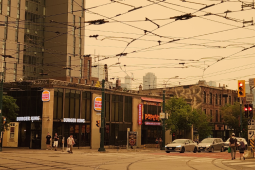 View of a city intersection. The sky, crisscrossed with power lines, is hazy grey and eerily tinted orange. Some people are out walking anyway.