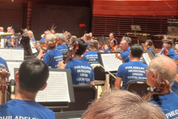 Orchestra musicians on a stage, apparently performing or rehearsing (conductor's baton is raised). Everyone, including the conductor, is wearing matching blue T-shirts that say "Philadelphia Musicians Union Local 77-274" on the back.