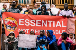 A diverse and serious crowd of people stands close together inside what looks like a church, in front of organ pipes, holding a big printed red banner that says "Essential not deportable" with the logo of Escucha Mi Voz. The person in the middle is speaking into a mic. Several kids sit in front and one holds a hand-lettered sign: "Free my grandma! Free her! I hate ICE!"