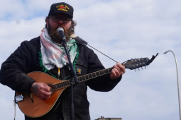 Emmett Doyle, a bearded white man wearing a "Class war veteran" hat, a keffiyeh, and a Carhartt jacket, plays an Irish bouzouki. He is outdoors, silhouetted against a blue sky with thin white clouds.