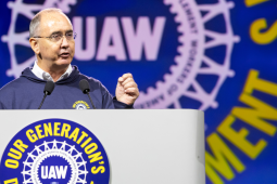 Shawn Fain, a white man with glasses, speaks at a podium. Behind him (and on the podium) is the UAW logo in white on a blue background. Yellow letters around it say (partly obscured) "Our Generation's Defining Moment."