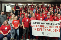 A group of dozens of teachers wearing red shirts gather behind a union banner.