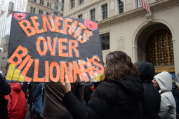 Marchers walk away from the camera with a giant orange on black sign, “Benefits over Billionaires”