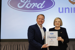 A man and a woman in suits stand holding a binder together in front of a screen that has Ford and Unifor logos projected on it.
