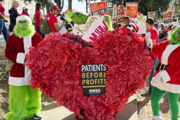 Two people in green Grinch suits hold a giant paper-mache heart that contains a "patients before profits" sign. Behind them an NUHW Kaiser picket line crowd is visible.