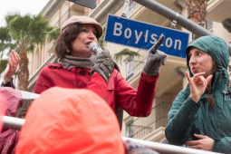 A woman speaks at a strike rally while another woman interprets in sign language. Behind them is a palm tree, an apartment buidling or hotel, and a street sign "Boylston"