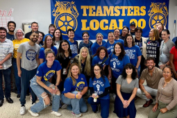 A group of 25 blue-shirted nurses pose happily in front of a Teamsters Local 90 banner