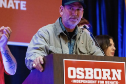 A man in a ball cap stands behind a lectern that says Osborn for Senate