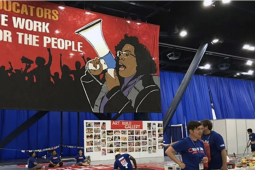 A banner says “Educators, we work for the people” in the background as union members are visible setting up for a meeting in the foreground. 