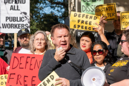 Tom Alter, a white man with a short haircut, squints into the sun and speaks into a megaphone, He is surrounded by a tightly packed crowd, diverse in race and gender, holding signs with slogans like "Defend Free Speech," "Defend All Public Workers," and "Reinstate Tom Alter."