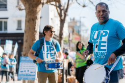 Two men in blue shirts that say “UPTE” play snare drums while others march in the background.