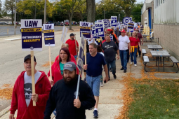 A dense line of picketers (at least 25 people are visible but it looks like there are more behind them) marches towards the camera on a leafy sidewalk alongside a factory. Many carry printed signs like "UAW on strike" and "Stand up for retirement security." Most visible faces are white but a few Black faces are also visible; there's a mix of women and men.