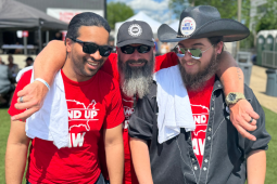 Three guys in red Stand-Up Strike (UAW) t-shirts, two bearded, stand outside on grass with arms around each other’s shoulders, smiling big.
