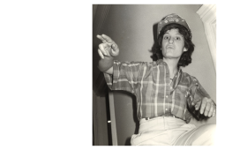 Black-and-white photo looks upward at a woman in a plaid shirt and bedazzled hardhat, gesturing and speaking