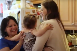 A white woman, right, hands a baby to a Latina woman, left. They are in an apartment kitchen. Both are smiling big. The baby's face is not visible.