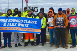 16 people stand facing the camera. Most are wearing jeans and neon yellow or orange jackets or hoodies. Several are helping to hold a blue banner: "Rehire Edward Clarke and Alex White, no retaliation or favoritism." Others hold signs: "180 hours paid time off, unionize KCVG" and "Unionize Amazon."