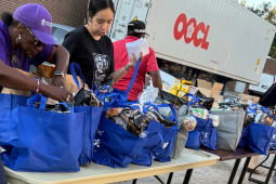 Three women work around a table with blue cloth grocery bags, outdoors. A man in a red T-shirt walks behind them. A truck is also visible.
