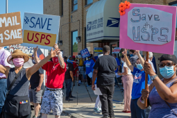 A group of people, masked, socially distanced, and diverse in race and gender, stand outside the Old Bedford Station post office on a sunny day. They hold up handmade signs: "US Mail Not 4 Sale" and "Save USPS." Someone in the back has a camera set up on a tripod. Similar printed signs from APWU are also visible.