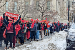 A long procession of strikers bundled up against the cold, most wearing bright red NYSNA hats or scarves, packs a sidewalk lined with piles of shoveled snow. The group is diverse in race and gender. Many are smiling. A woman in the foreground holds up a red "In Solidarity" banner.