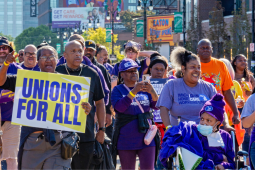 Workers march through a Detroit street, part of a Labor Day march. The people visible are mostly wearing purple SEIU T-shirts, many are Black and one holds a big printed sign readgin "Unions for all" on a yellow background. Another printed sign says "Families first, not billionaires. Not one more cut." One woman in a "Michigan home care workers" shirt pushes another in a wheelchair, also in purple gear.
