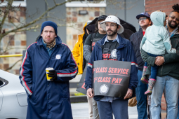 A racially diverse, young, mostly male crowd stands in front of a post office. Some people wear postal gear, others wear "Fight Like Hell" logo T-shirts, and some hold printed "First class service, first class pay" signs. One man holds a toddler in his arms. People wear coats and hats, and one open umbrella is visible in the back.
