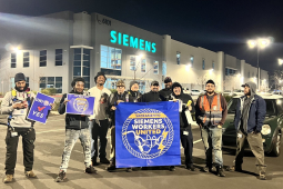 Siemens workers pose in front of the factory in the dark behind a blue banner that says Siemens Workers United.