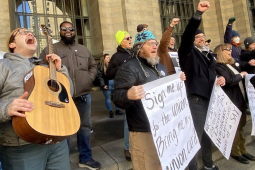 A group of people stands outside a building, singing. Edwin, a young man with guitar, has his head leaned back shouting into the sky. Several other people have fists in the air, one wears a big smile, and some hold big paper on which lyrics are written out: "Sign me up for the union, bring me my union card, We can work better with decent pay, health care, childcare, and more of a say..."