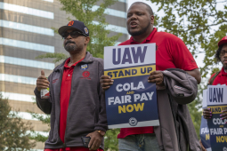 A group of workers march in Detroit holding signs that say "UAW Stand Up: Fair Pay and COLA Now." 