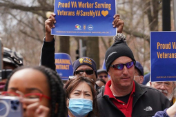 several people are jammed together as part of a march with blue signs with gold lettering, saying “Proud VA Worker Serving Veterans, #SaveMyVA”