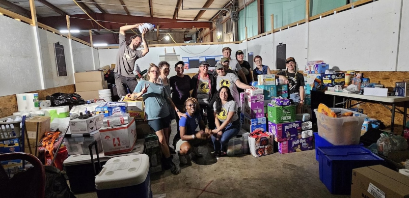 A group of fifteen workers pose inside a warehouse among boxes of diapers and other aid material