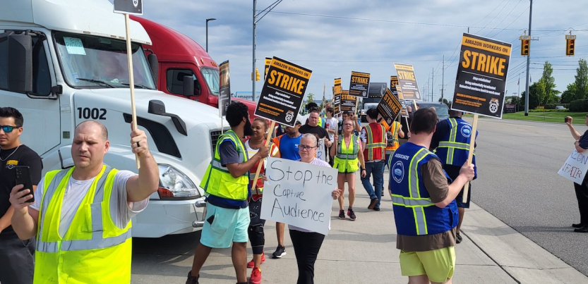 Workers in high-visibility vests picket with ‘Amazon workers on strike’ signs picket by a road, one handmade sign reads ‘Stop the captive audience’