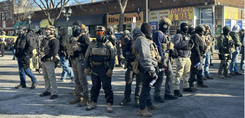 Agents covered head to toe in tactical gear and warm clothing stand in a diamond formation, facing outward, in a city street. Their faces are covered. The vests say "police" though they are not in uniform. They all wear heavy boots and some carry spray cans. Visible behind them is a one-story brick building lined with retail shops, with faded signs reading "8 Street Nourish" and "Tobacco & Mas."