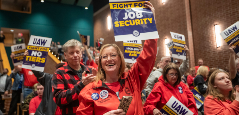 People, mostly wearing red, hoist printed signs in a packed indoor rally. A woman in the foreground, smiling, wears a "no tiers" button. Her sign, held high, reads "UAW: Fight for Job Security." Other visible signs say "UAW: Fight for No Tiers" and "UAW: It's Our Time at Local 2093 American Axle." A man behind her, applauding, wears a red and black buffalo plaid shirt. Most people visible in the photo appear to be white.