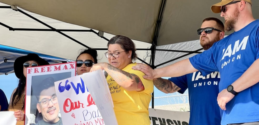 A woman cries while two men in blue Machinists t-shirts comfort her with a hand on her shoulder. Next to her is a woman holding a sign that says “Free Max.”