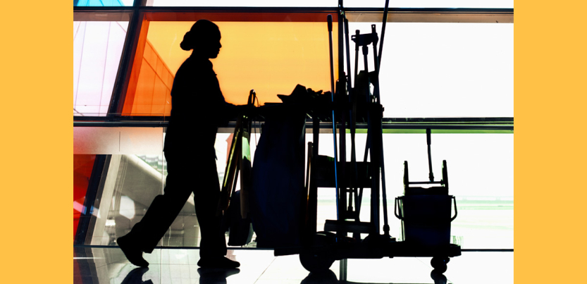 A woman custodian pushes a cart full of implements in profile past colorful windows.