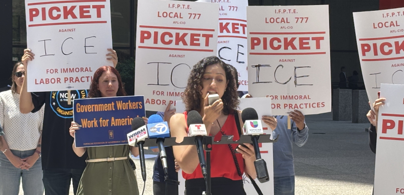A woman speaks into a microphone outside while others stand behind her with signs that say “Picket ICE”