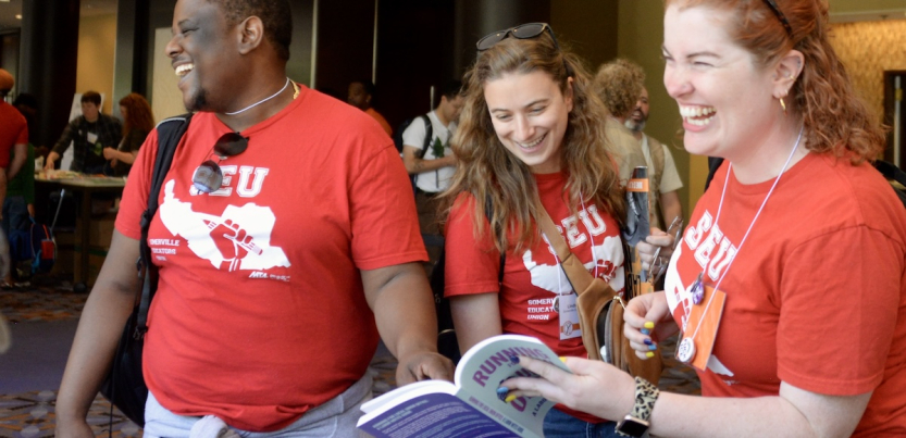 Two women and a man, all wearing Somerville Educators red T-shirts, laugh as they look at a book