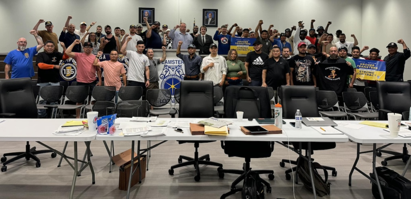 A group of 50 people stand behind a bargaining table with fists raised. 