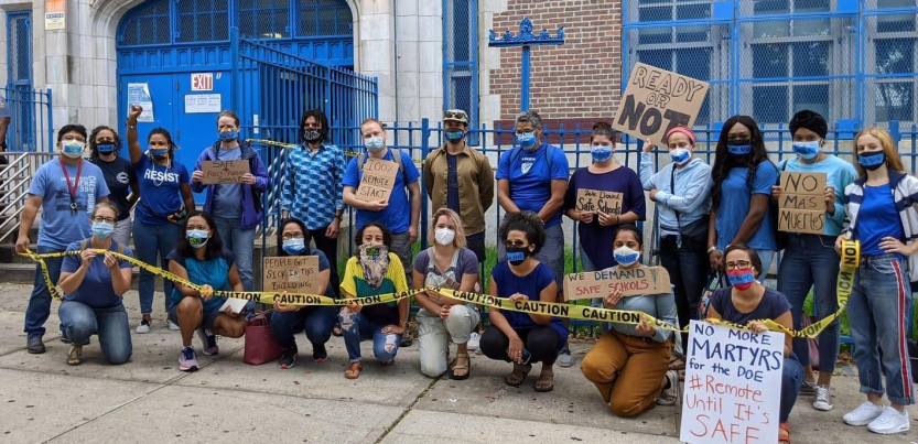 21 adults in masks outside a school. Together they hold "CAUTION" tape. Handmade signs say "100% remote start," "no mas muertes," "ready or not," "we demand safe schools," "no more martyrs for the DOE #Remote until it's safe" and "people got sick in this buidling."